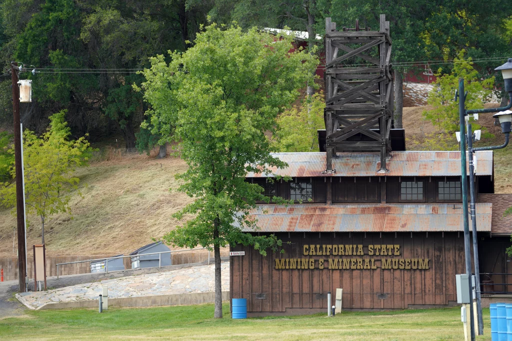 Außenansicht des California State Mining & Mineral Museum mit rostigem Blechdach und altem Förderturm, aufgenommen in Mariposa, Kalifornien.