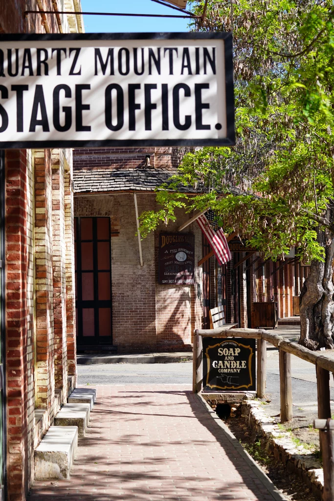 Historische Backsteinfassaden und Ladenschilder in Columbia, Kalifornien, mit dem Schild „Quartz Mountain Stage Office“.