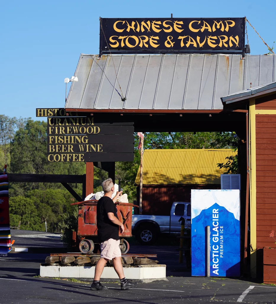 Mann vor dem Schild des „Chinese Camp Store & Tavern“ mit Eisautomat, Erz-Lore und historischen Hinweisen