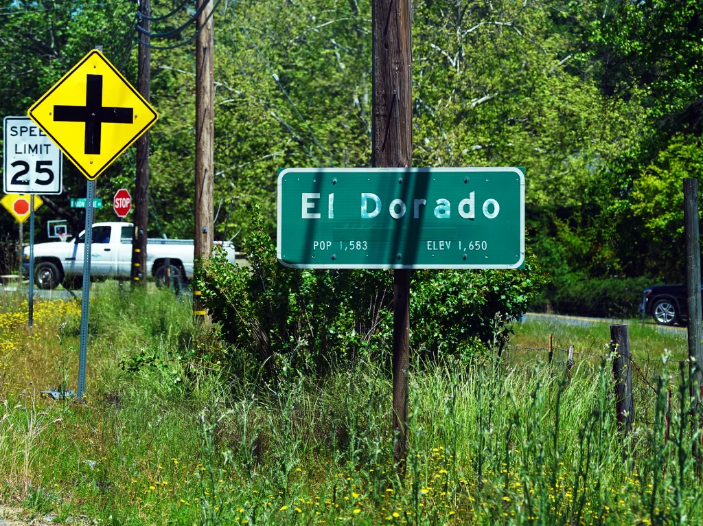 Ortsschild von El Dorado, Kalifornien, an einer Landstraße mit Verkehrsschildern im Hintergrund