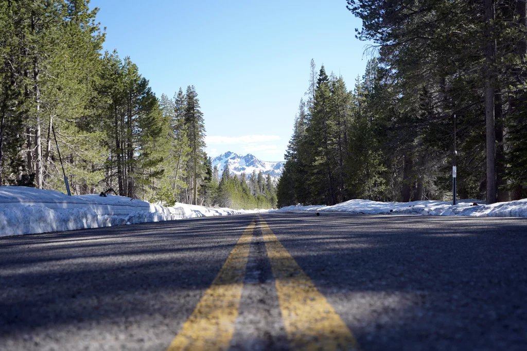 Blick auf eine verschneite Straße durch Wald mit schneebedeckten Bergen im Hintergrund, aufgenommen auf dem Highway 49 bei Sierra City, Kalifornien.
