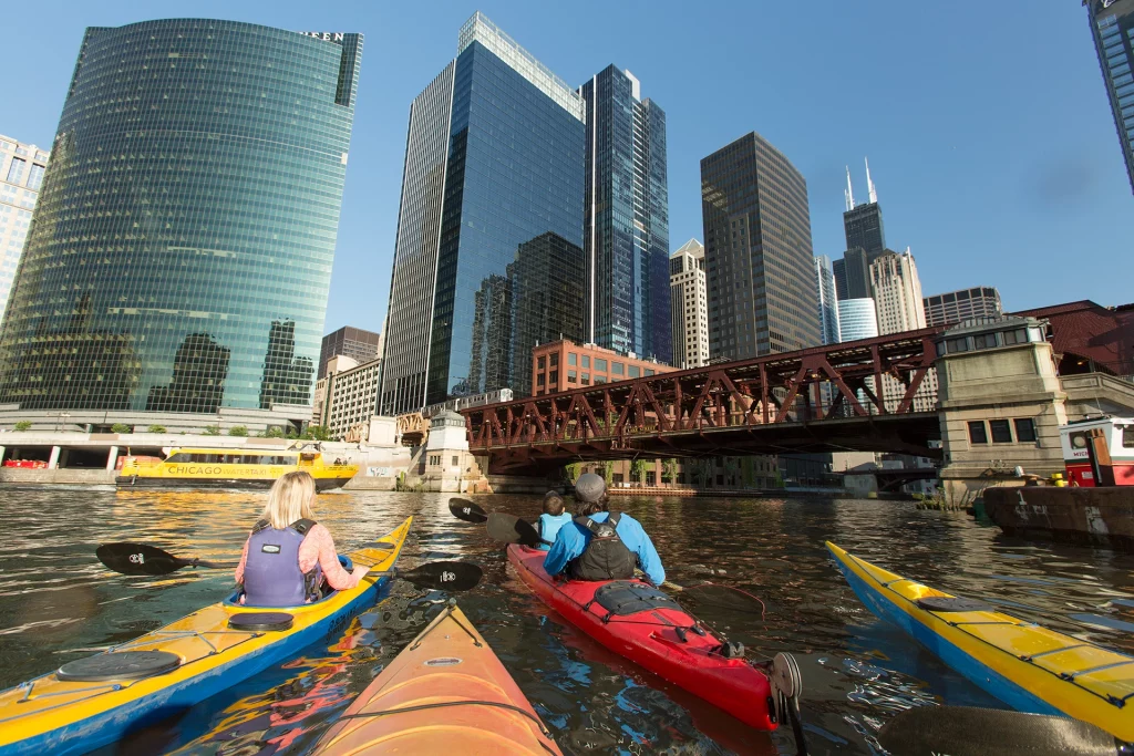 Kajakfahrer paddeln auf dem Chicago River mit Blick auf die Skyline und eine historische Eisenbahnbrücke.