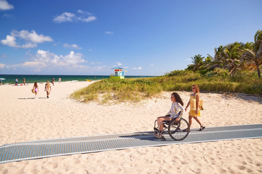Frau im Rollstuhl auf barrierefreiem Strandweg in Fort Lauderdale, begleitet von einer Freundin.
