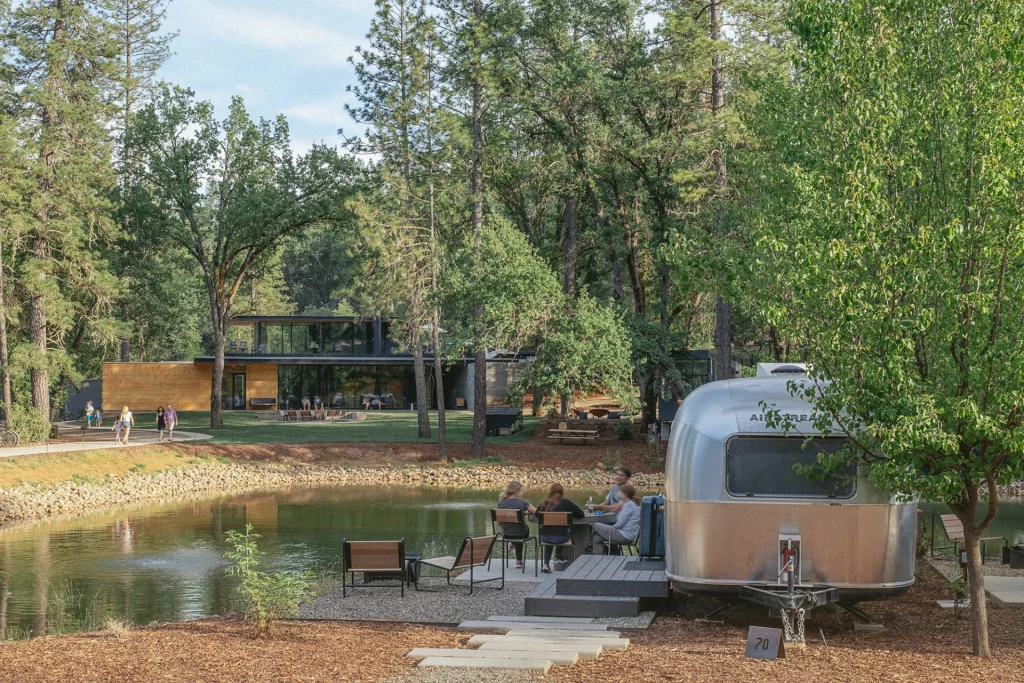 Moderner Airstream-Wohnwagen am Teich im AutoCamp Yosemite, umgeben von Bäumen und Gästen im Freien.