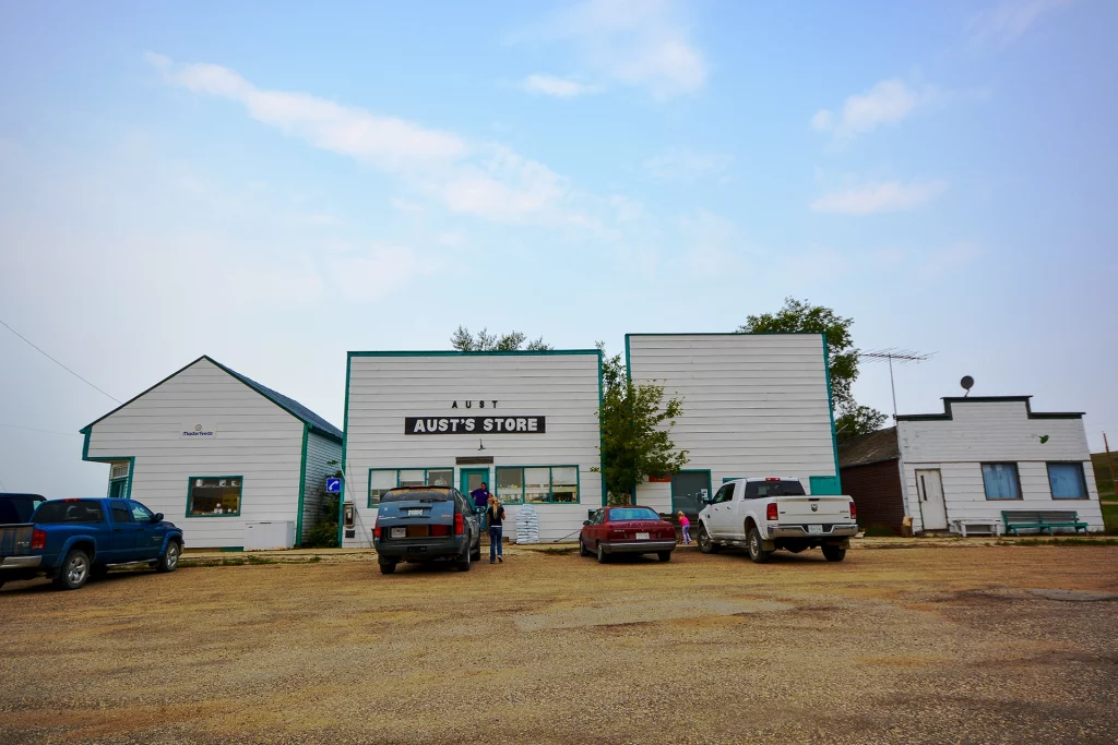Fassade von Aust’s Store in Big Beaver, Saskatchewan, mit angrenzenden Gebäuden und parkenden Autos auf einem Schotterparkplatz.