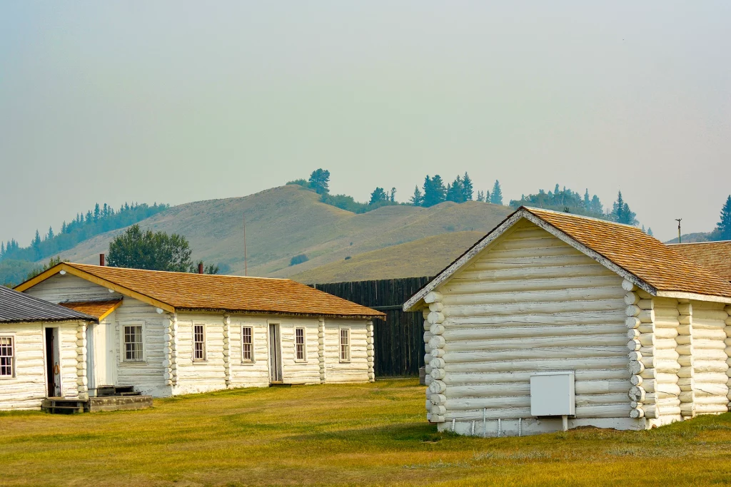 Restored log buildings of Fort Walsh in Saskatchewan surrounded by rolling hills and pine trees.