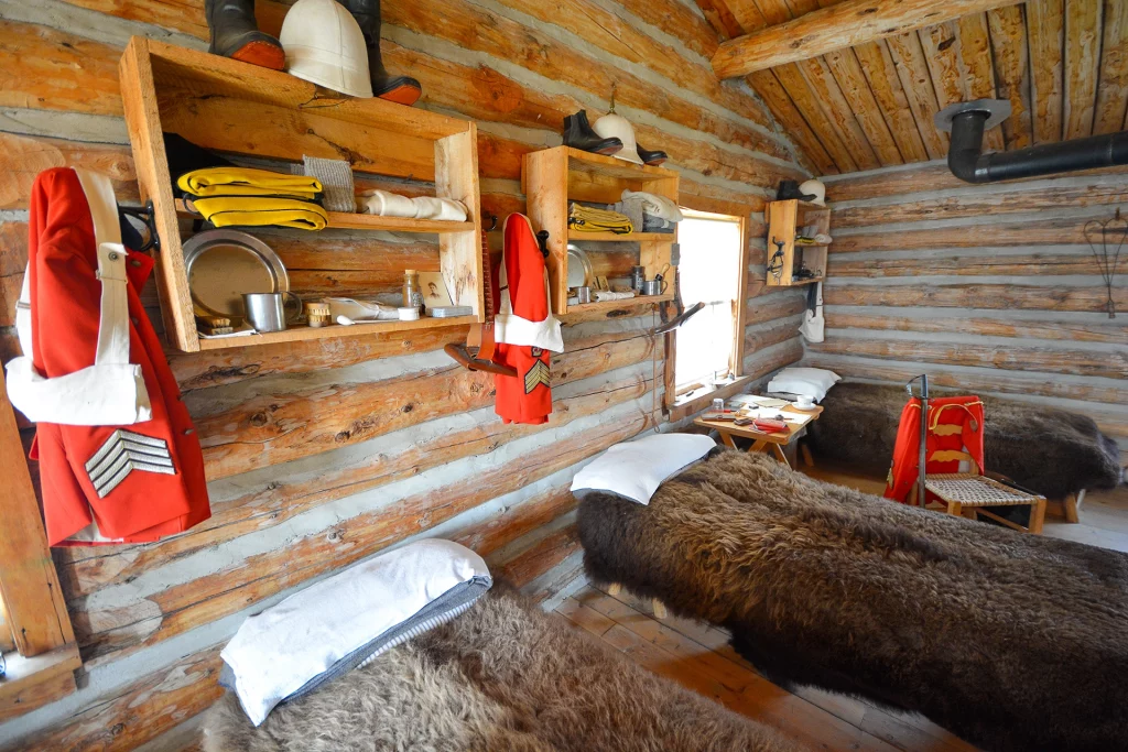 Interior of Fort Walsh showing bunk beds, buffalo hides and red NWMP uniforms on log walls.