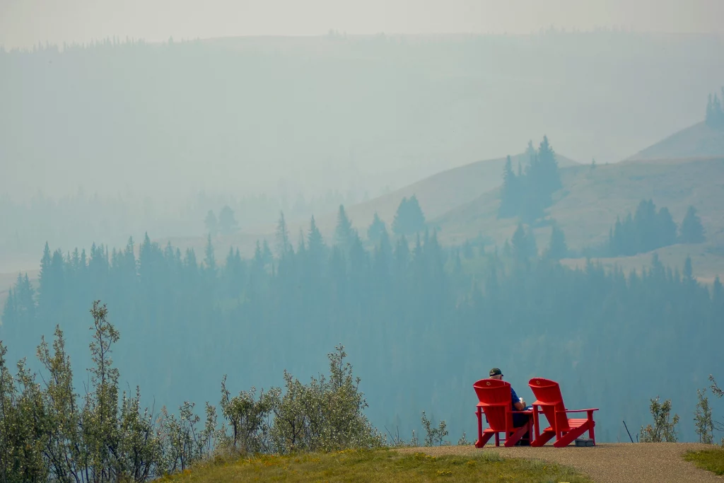 Zwei rote Adirondack-Stühle mit Person vor rauchverhangenem Himmel und bewaldeten Hügeln in Fort Walsh, Saskatchewan