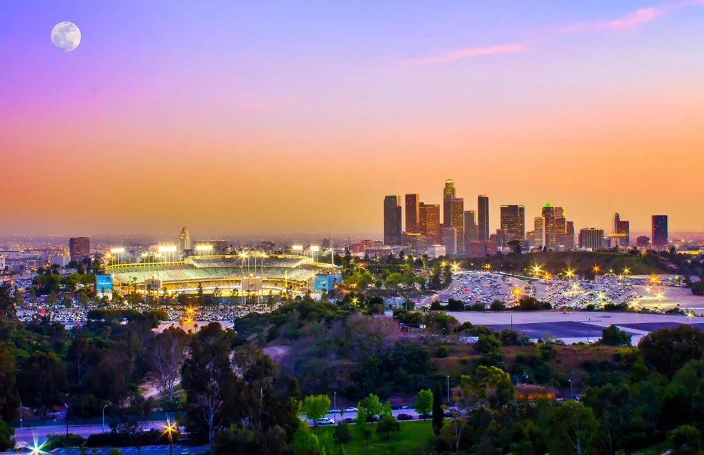 Dodger Stadium illuminated at sunset with downtown Los Angeles skyline in the background.