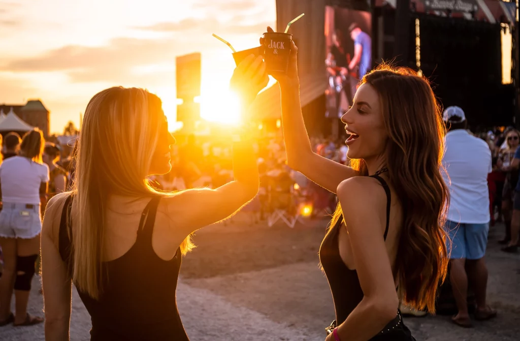 Zwei junge Frauen stoßen bei Sonnenuntergang auf einem Musikfestival in Louisville an.