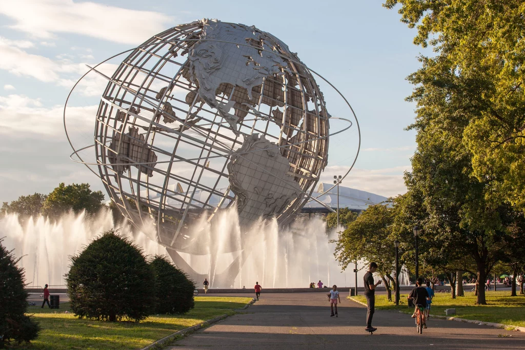 Metallene Unisphere-Skulptur mit Wasserfontänen im Flushing Meadows Corona Park in Queens, New York, umgeben von Spaziergängern und Radfahrern.