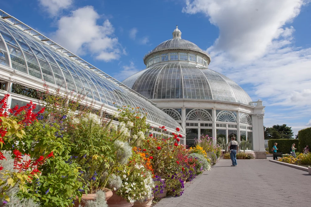 Glashaus des New York Botanical Garden in der Bronx, umgeben von bunten Blumenbeeten unter blauem Himmel.
