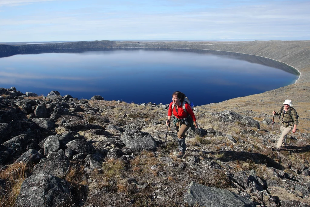 Zwei Wanderer mit Trekkingausrüstung steigen über felsiges Gelände am Rand des tiefblauen Pingualuit-Kratersees in Nunavik, Québec.