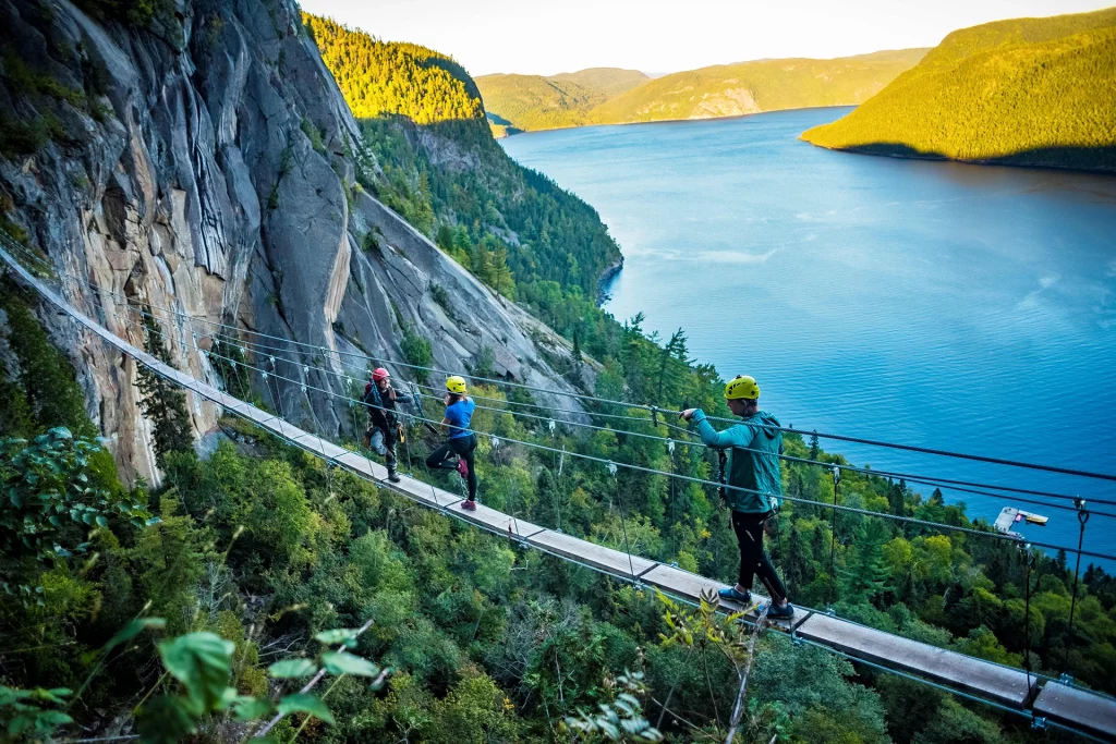 Drei Kletterer mit Helmen überqueren eine Hängebrücke an einer steilen Felswand über dem Fjord Saguenay in Québec.