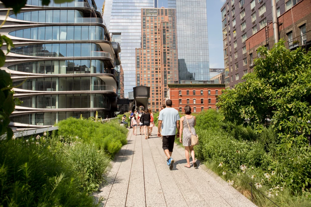 Spaziergänger auf der begrünten High Line in Chelsea, flankiert von moderner Glasarchitektur und Blick auf die Skyline.