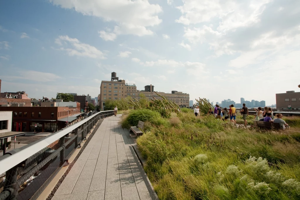 Besucher spazieren entlang der High Line in Manhattan, flankiert von üppiger Vegetation und Blick auf die umliegende Stadt.