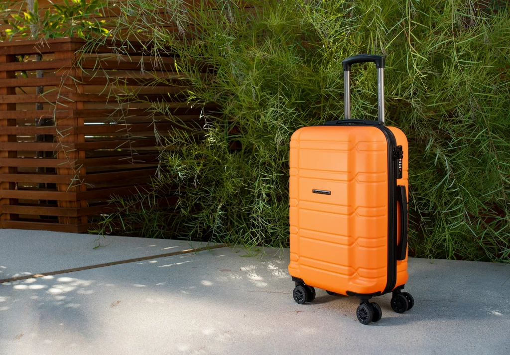 Orangefarbener Hartschalenkoffer mit Teleskopgriff und vier Rollen, stehend auf einer Terrasse vor grünen Pflanzen.