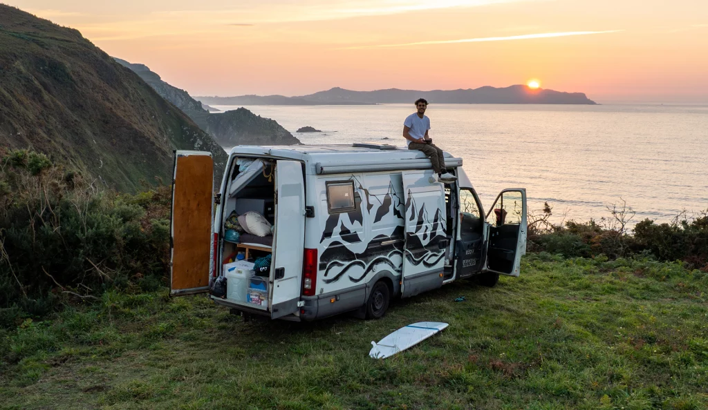 Mann sitzt auf einem selbst ausgebauten Van bei Sonnenuntergang an der Steilküste