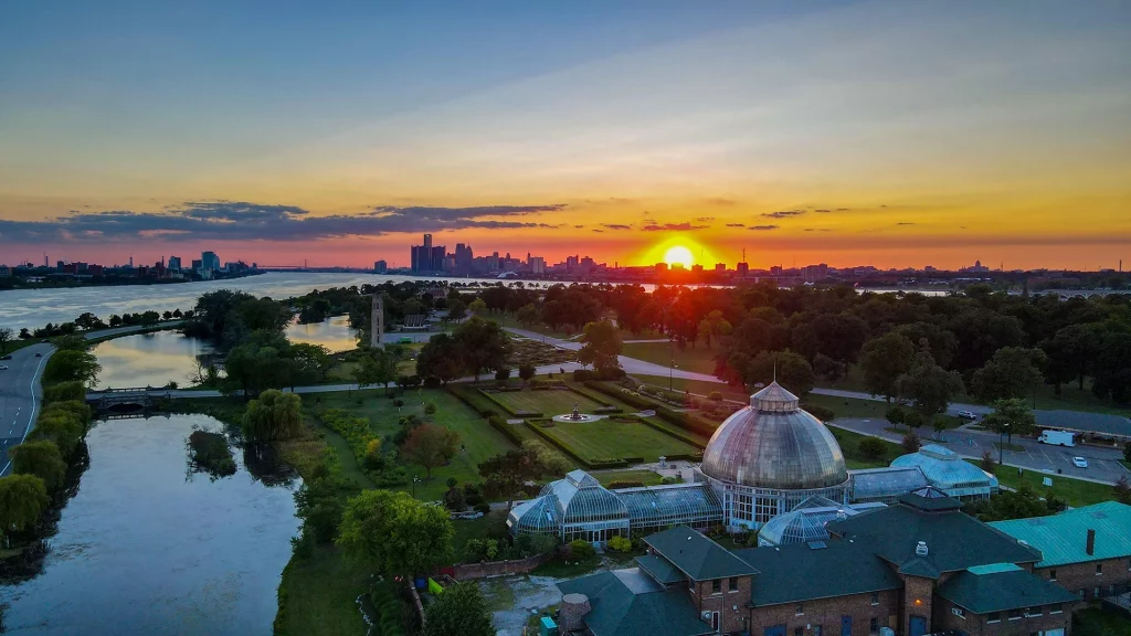Luftaufnahme von Belle Isle mit dem Anna Scripps Whitcomb Conservatory im Vordergrund und der Skyline von Detroit bei Sonnenuntergang.