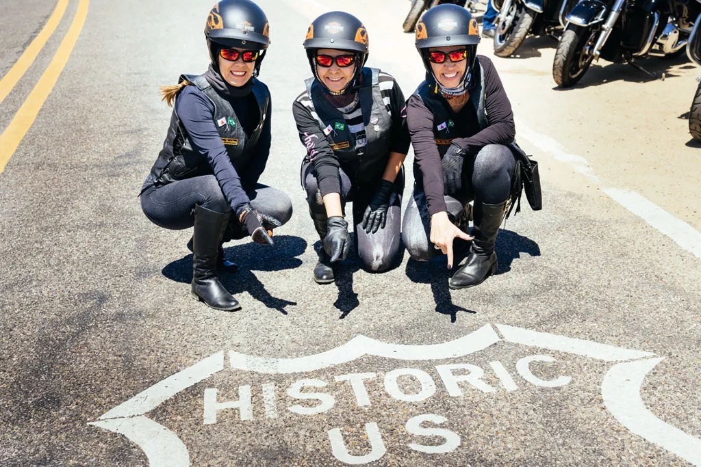 Drei Motorradfahrerinnen posieren am Historic Route 66 Schild auf dem Asphalt.