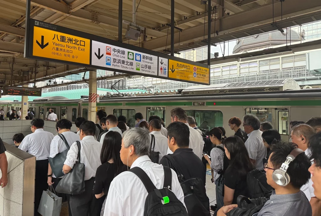 Menschenmenge am Bahnsteig in Tokio mit Yamanote-Linienzug im Hintergrund.