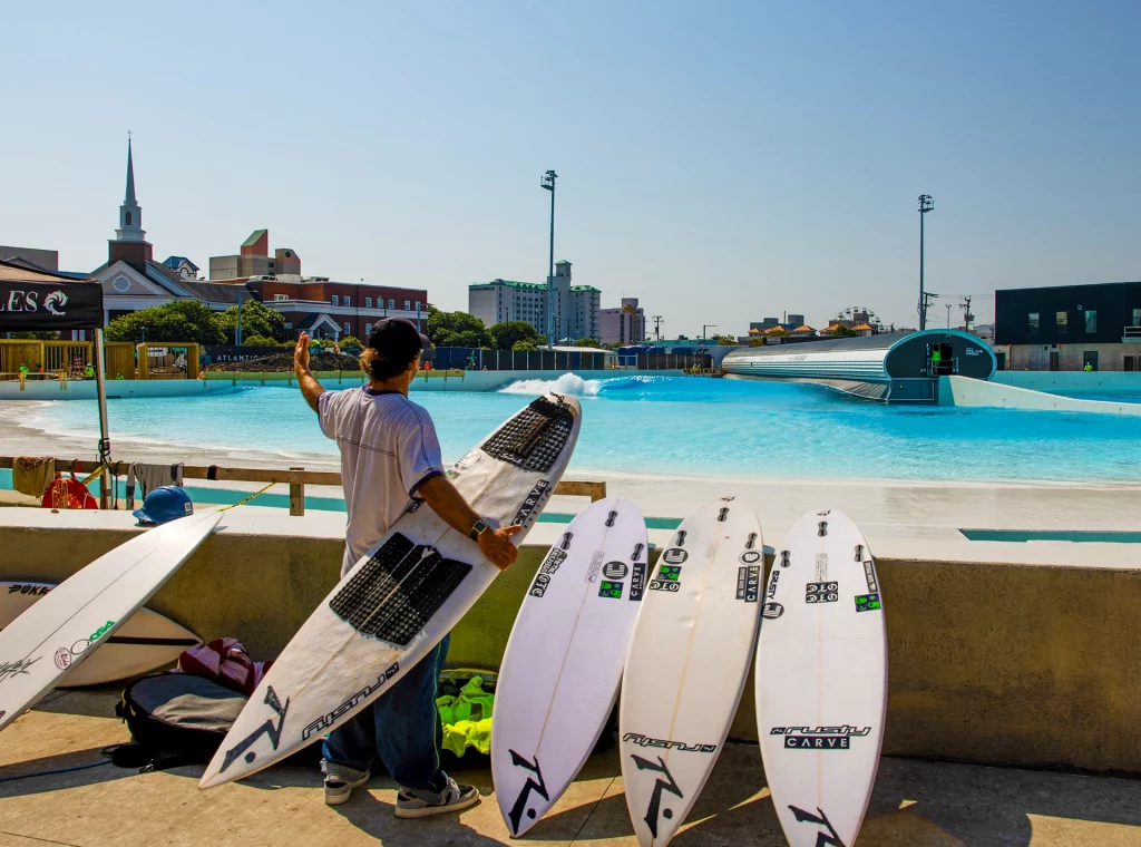 Ein Surfer mit mehreren Boards steht am Beckenrand des Atlantic Park Surf Wavepools in Virginia Beach und beobachtet die erzeugten Wellen.