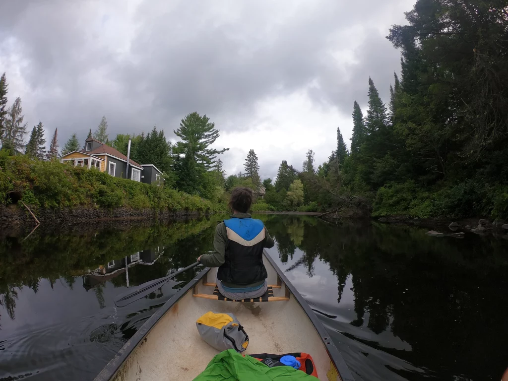 Person paddelt im Kanu auf einem ruhigen Fluss bei Tremblant, umgeben von dichten Wäldern und Ferienhäusern.