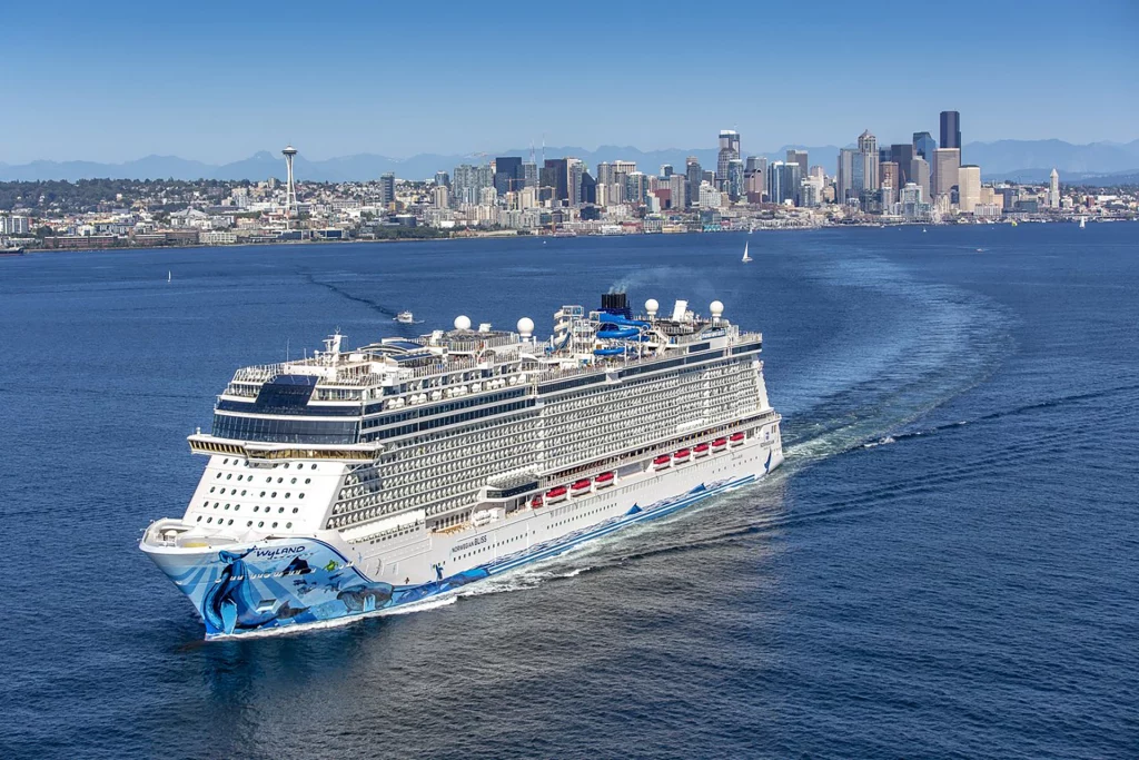 Das Kreuzfahrtschiff Norwegian Bliss verlässt den Hafen von Seattle mit Skyline im Hintergrund.