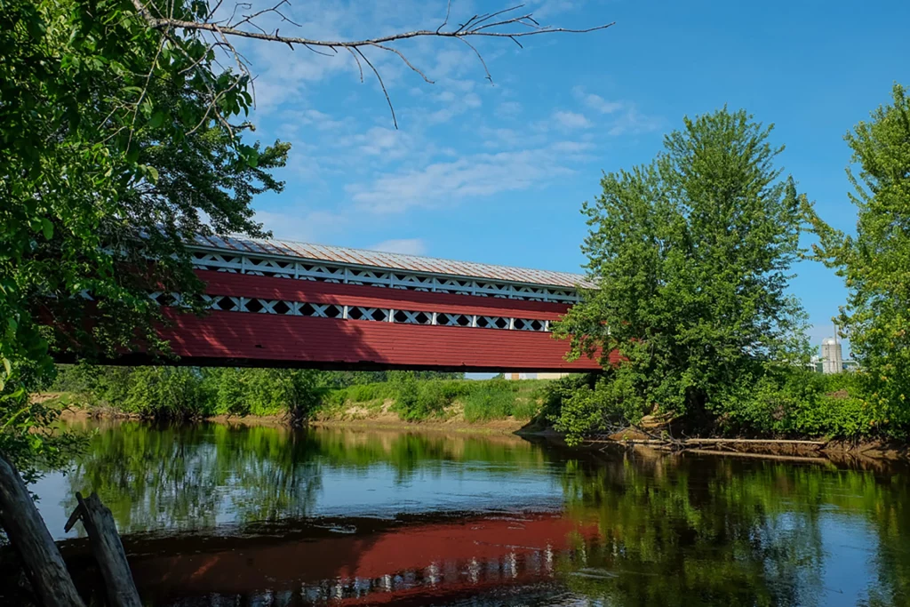 Historische rote Holzbrücke Pont Prud’homme über einem Fluss in den Laurentiden, Québec, mit Spiegelung im Wasser.