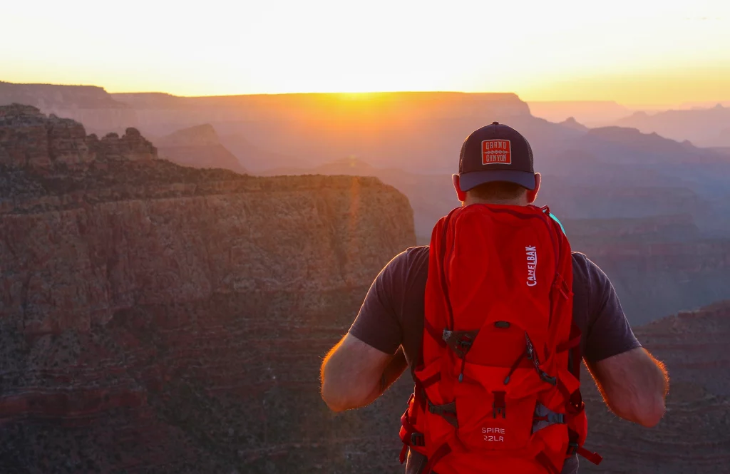 Wanderer mit rotem Rucksack blickt bei Sonnenaufgang über die Schluchten des Grand Canyon.