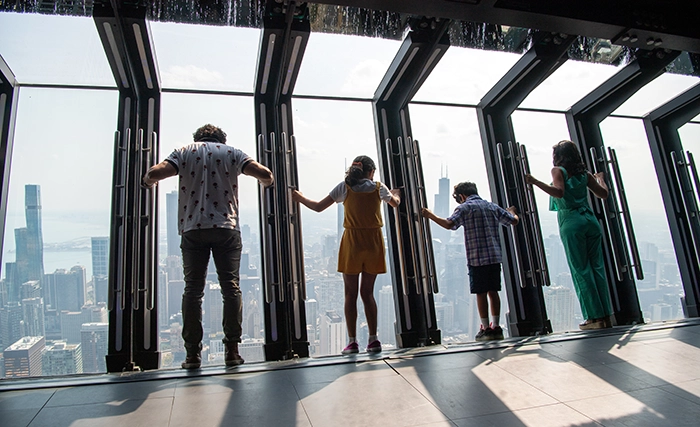 Besucher lehnen sich auf der gläsernen Plattform „Tilt“ im 360 Chicago Observation Deck über den Rand des John Hancock Centers und blicken spektakulär auf die Skyline.