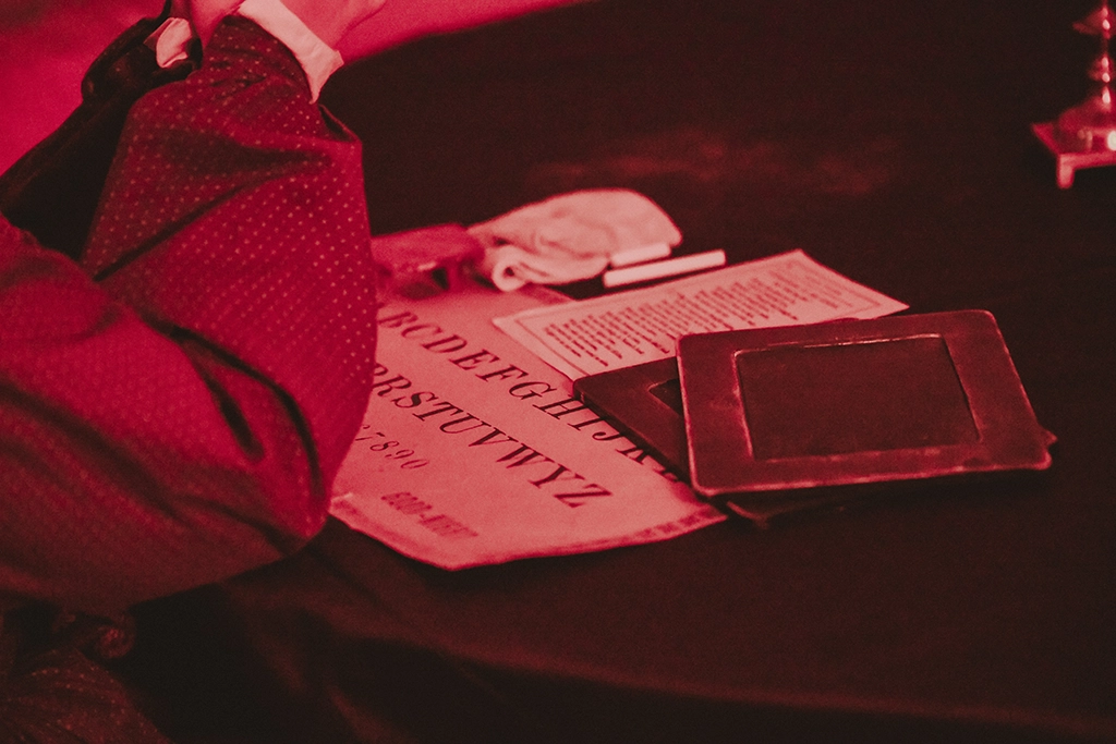 Eine Person sitzt an einem Tisch mit einer alten Ouija-Tafel und Kerzenlicht in rotem Schein.