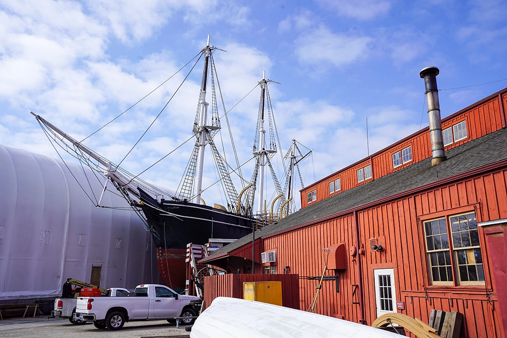Ein historisches Segelschiff im Mystic Seaport Museum in Connecticut, umgeben von roten Werfthallen und maritimen Werkstätten unter blauem Himmel.