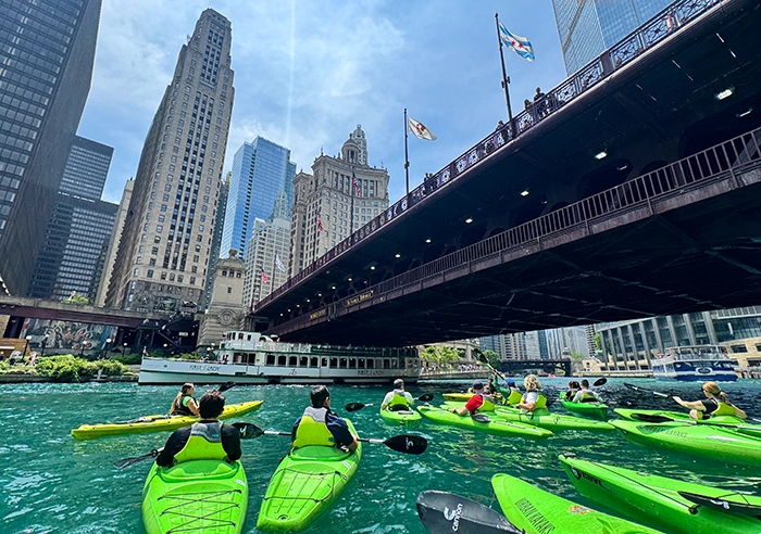 Menschen paddeln in grünen Kajaks auf dem Chicago River unter einer Brücke, umgeben von Hochhäusern in der Innenstadt von Chicago.