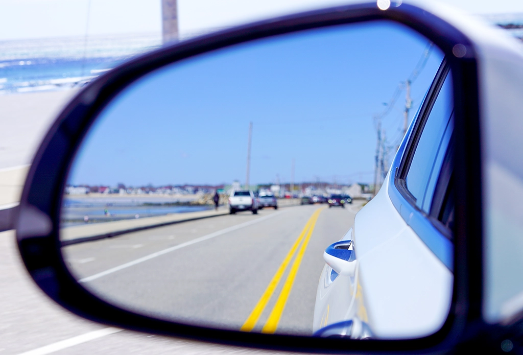 Blick in den Rückspiegel eines Autos auf eine Küstenstraße in Neuengland mit blauem Himmel und Meer im Hintergrund.