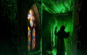 Eine unheimliche, grün erleuchtete Szene mit einer dunklen Gestalt in Mönchskutte vor einem Kirchenfenster.