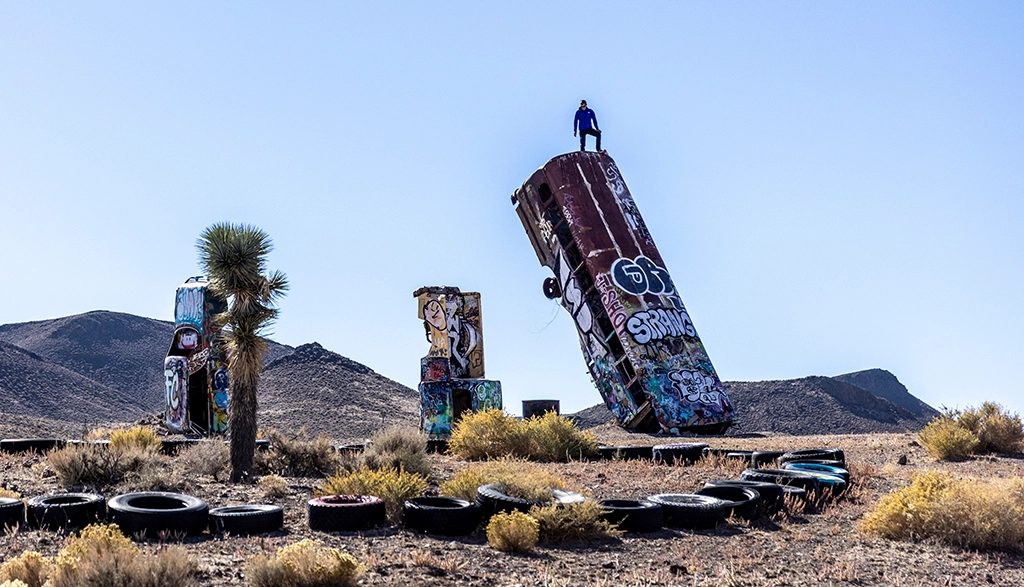 Ein Mann steht auf einem senkrecht im Boden steckenden, bunt bemalten Autowrack in der Wüste von Nevada.