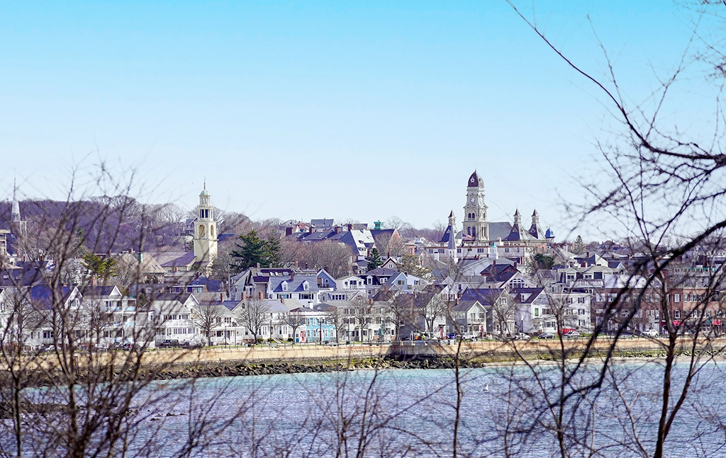 Blick auf die historische Altstadt und Hafenfront von Gloucester, Massachusetts, mit Kirchtürmen und Häusern entlang der Küste.