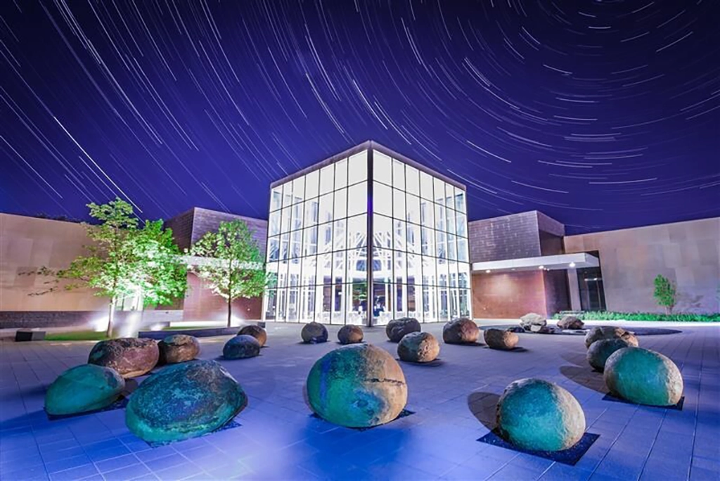 Glaspavillon des North Dakota Heritage Center & State Museum bei Nacht, umgeben von Felsblöcken und Sternenspuren am Himmel.