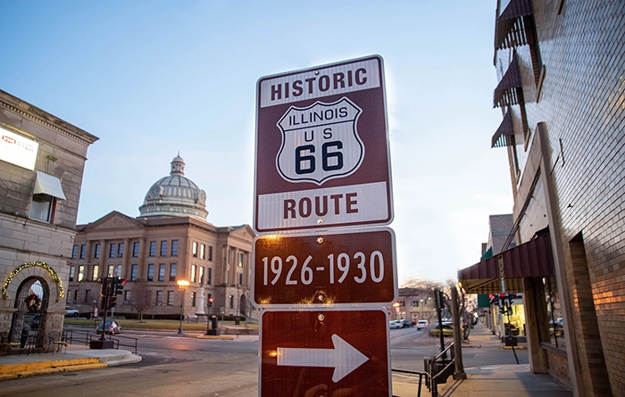 Straßenschild der historischen Route 66 in Illinois mit Aufschrift „1926–1930“, im Hintergrund die Altstadt von Lincoln, Illinois, bei Abendlicht.