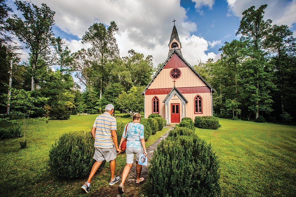 Ein älteres Paar geht auf einem gepflegten Pfad auf eine kleine, historische Holzkirche im Wald zu.