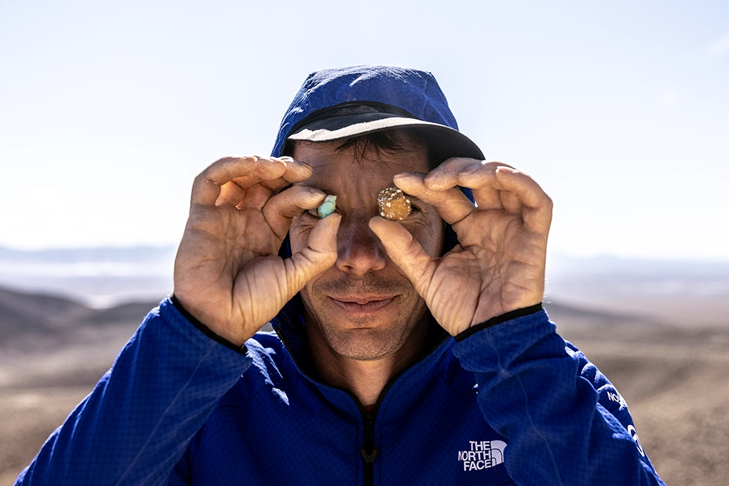 Alex Honnold hält zwei kleine Mineralsteine vor seine Augen und blickt durch sie in die Kamera.