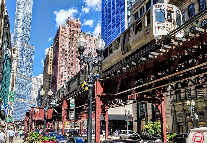 Ein Zug der Chicago „L“ Hochbahn fährt auf der charakteristischen Stahltrasse durch die Innenstadt, umgeben von Wolkenkratzern und Stadtverkehr.
