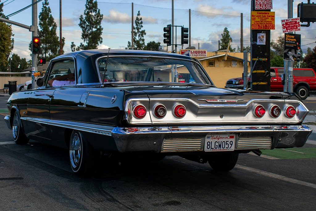 Schwarzer Chevrolet Impala mit kalifornischem Kennzeichen steht an einer Kreuzung unter Palmen und Neonlichtern.