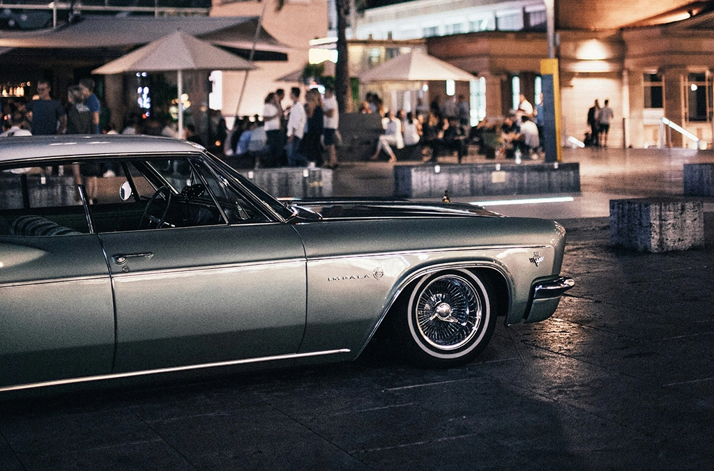 Seitliche Ansicht eines hellgrünen Chevrolet Impala bei Nacht, geparkt vor einer belebten Straßenszene.