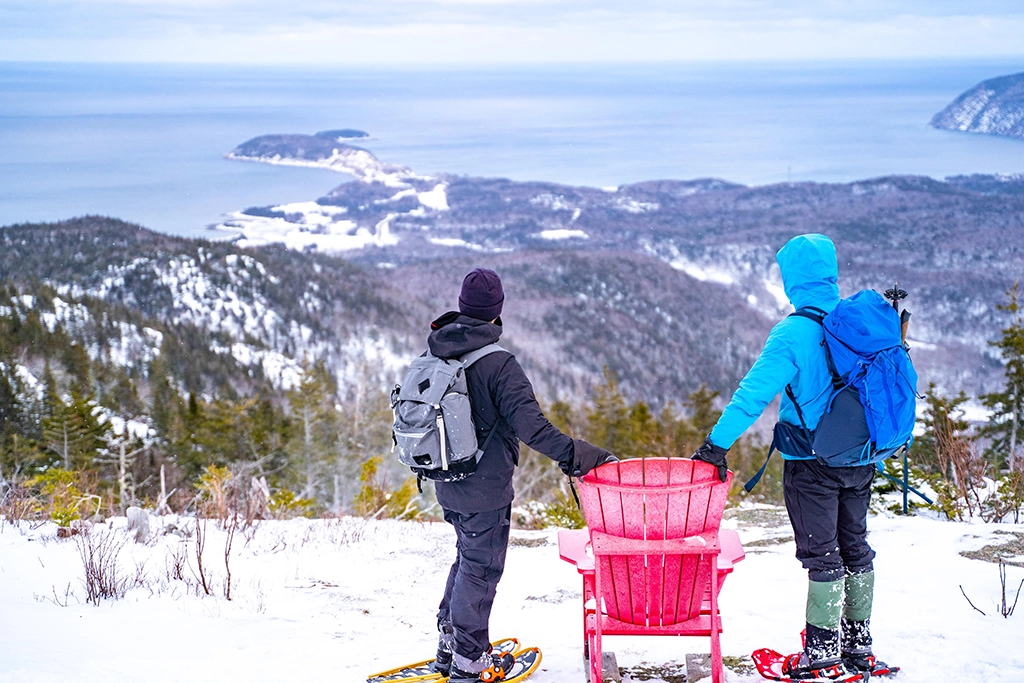 Zwei Schneeschuhwanderer blicken von einem Aussichtspunkt mit rotem Parks-Canada-Stuhl über den verschneiten Cape-Breton-Highlands-Nationalpark bis hin zur Küste.