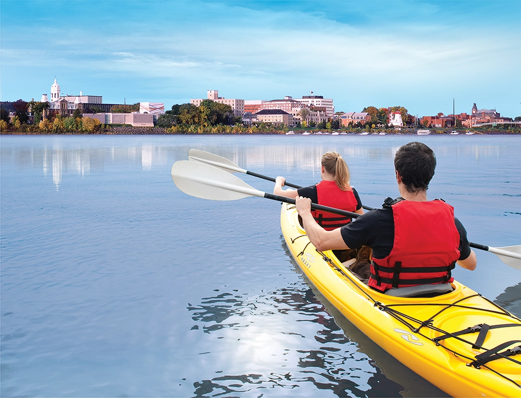 Zwei Personen paddeln im Kajak auf dem Saint John River mit Blick auf die Skyline von Fredericton, New Brunswick