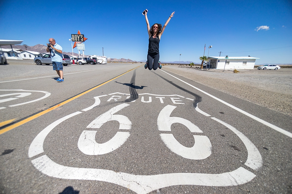Frau springt auf der Straße über das ikonische Route-66-Logo vor dem historischen Roy’s Motel Café in der Mojave-Wüste