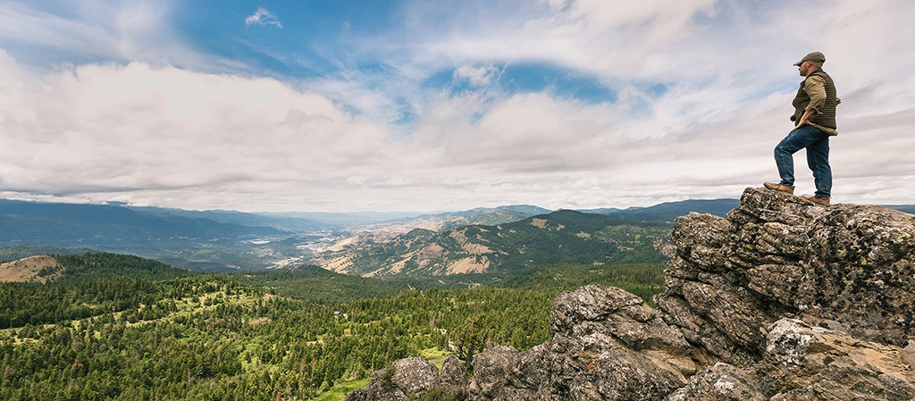 Ein Mann steht auf einem Felsen mit weitem Blick über die bewaldeten Berge Süd-Oregons unter dramatischem Himmel.