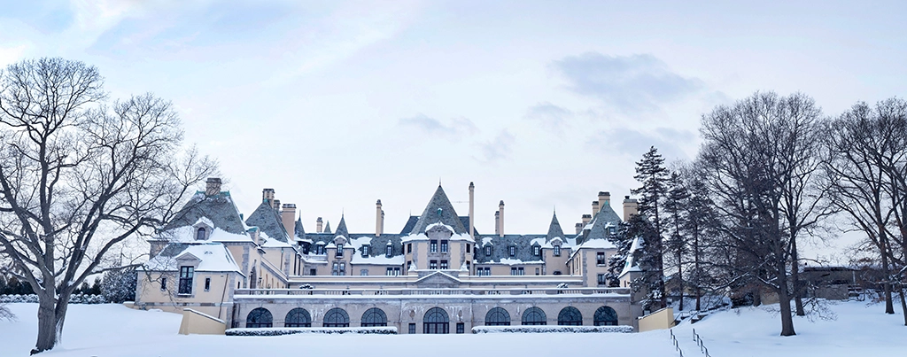 Winterliche Frontansicht des Oheka Castle mit verschneitem Rasen und Bäumen unter grauem Himmel